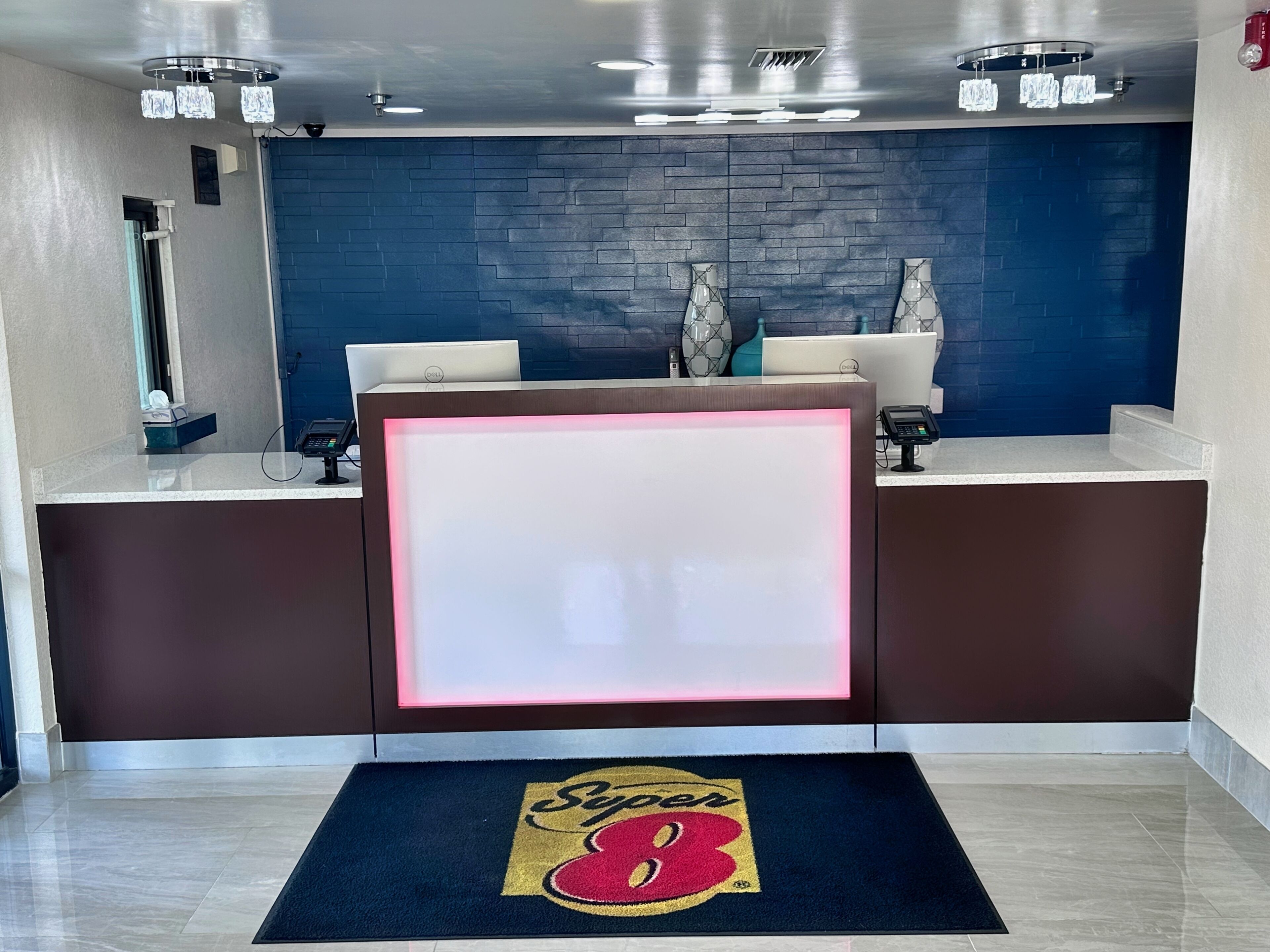 A hotel reception area with a modern design, featuring a reception desk, two computer monitors, a cash register, and a digital screen.