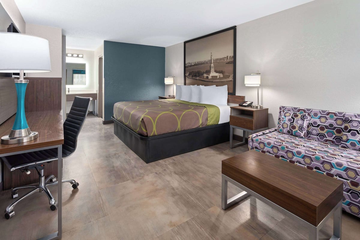 A modern hotel room with a bed, desk, chair, sofa, and a painting on the wall.