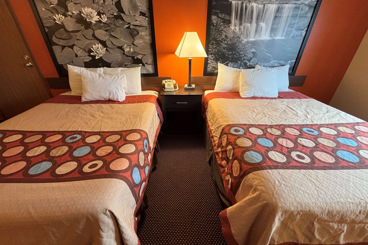 A hotel room with two beds, each with a patterned bedspread, a nightstand with a lamp, a telephone, and a wall with a waterfall painting.