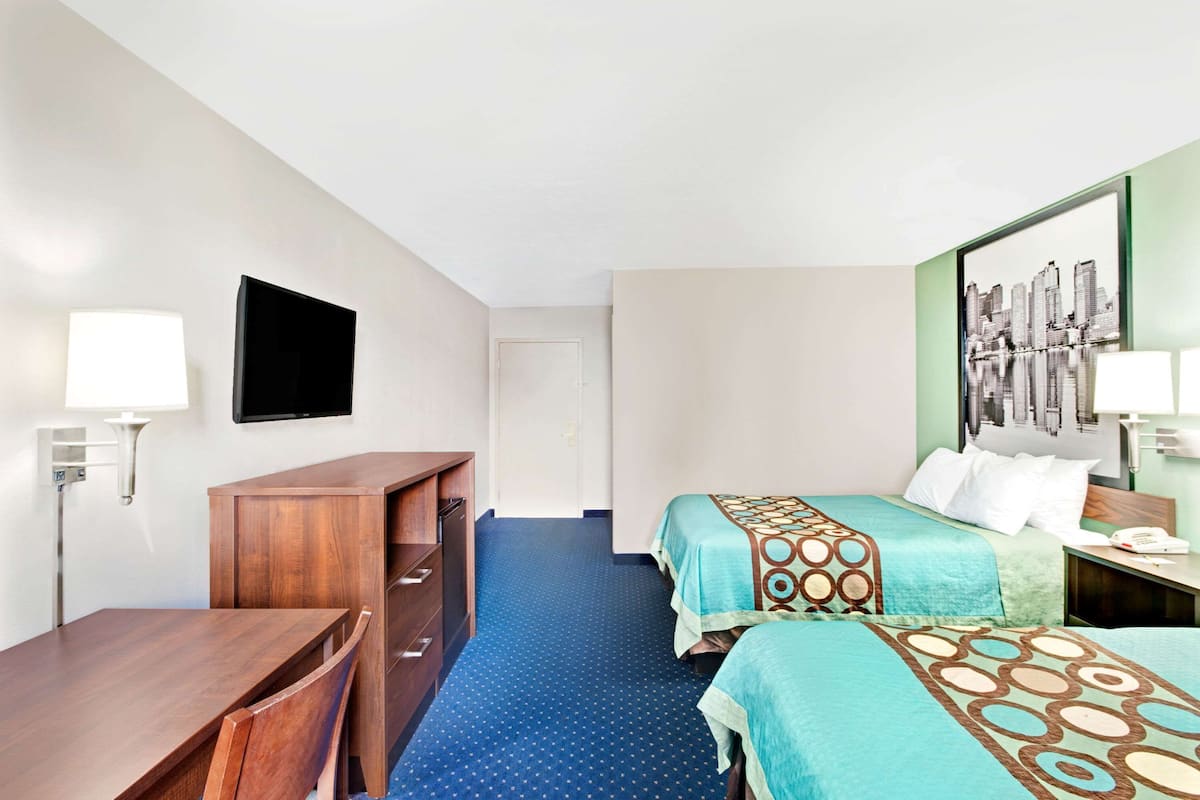 Une chambre d’hôtel avec un bureau en bois, deux lits recouverts de couvre-lits à motifs, un téléviseur à écran plat et une œuvre d’art représentant un paysage urbain accrochée au mur.