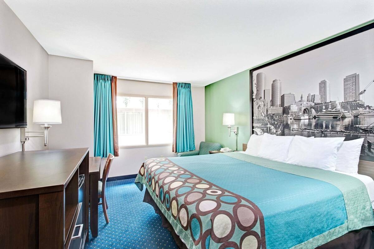 Une chambre d’hôtel comprenant un lit, un bureau, une chaise, une fenêtre avec des rideaux, un téléviseur fixé au mur et une fresque représentant un paysage urbain.