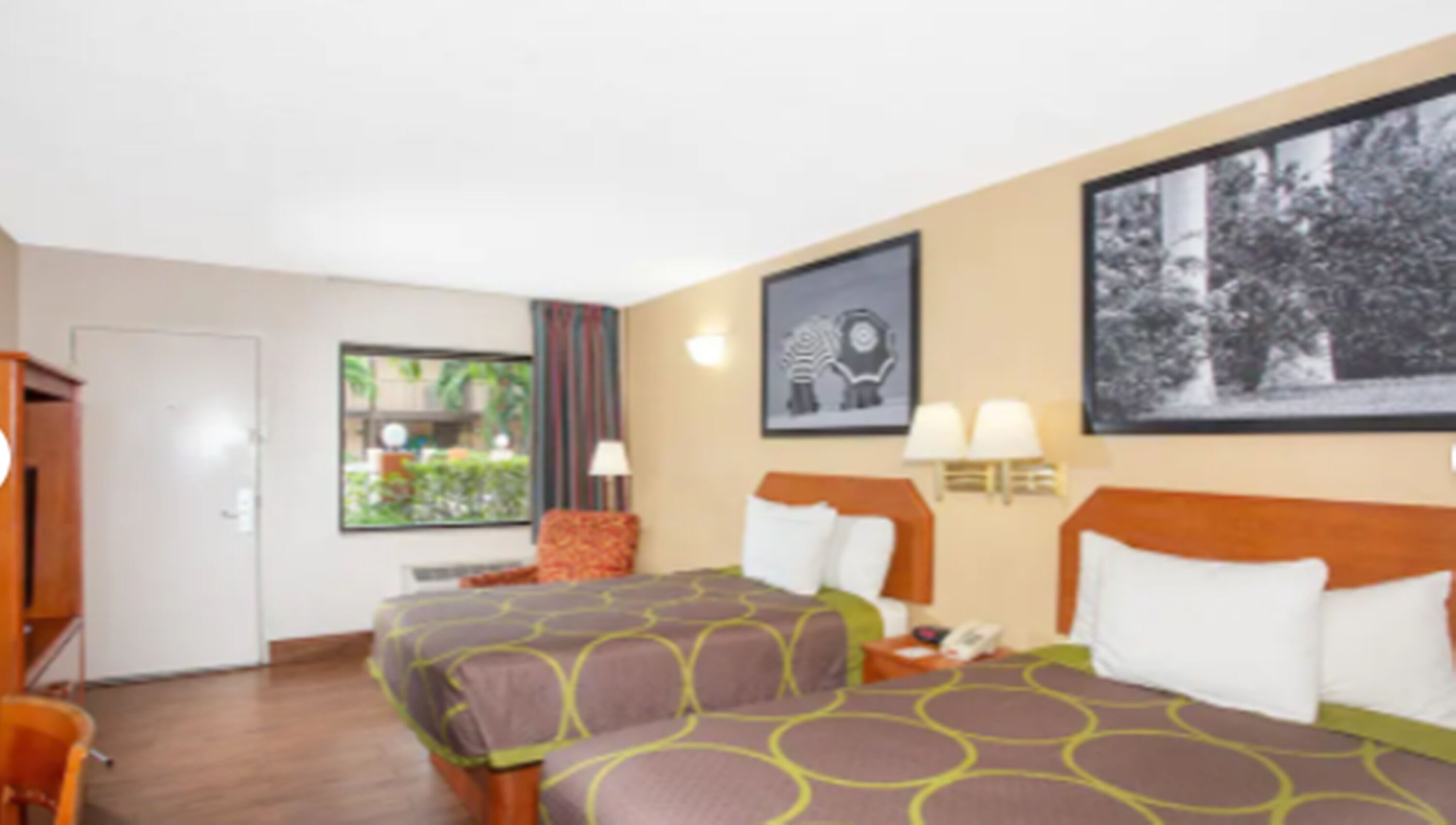 A hotel room with two beds, a desk, a chair, a window with a view of greenery, and artwork on the walls.