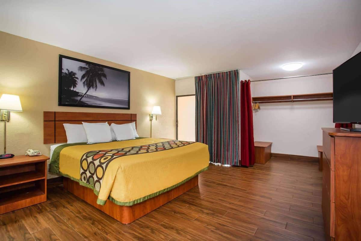 A hotel room with a bed, bedside lamps, a TV, a wooden desk, a coat rack, and a painting on the wall.