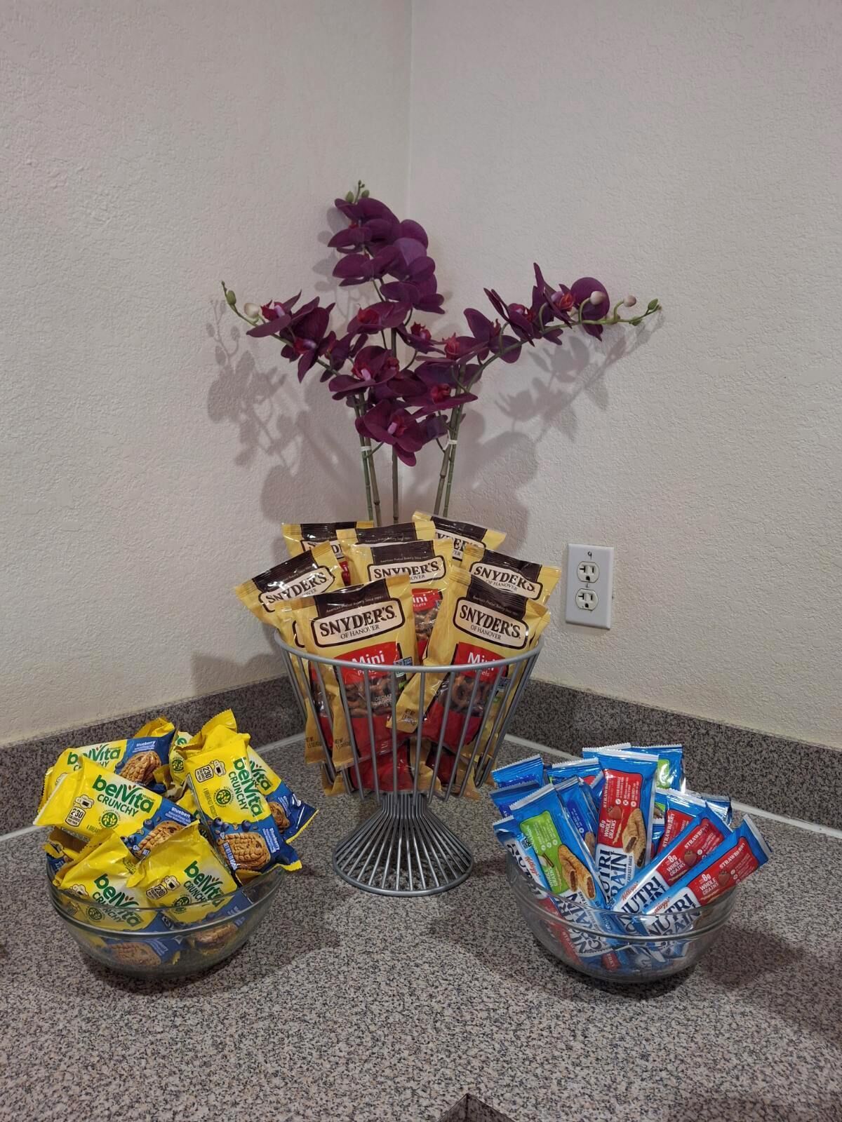 Una presentación de bocadillos con bolsas de Belvita Crunchy y pretzels Mini Snyder's, rodeada por un arreglo decorativo de orquídeas moradas.