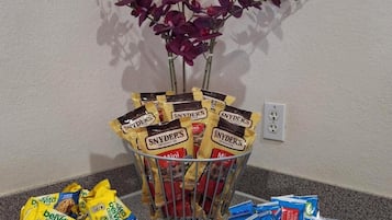 Una presentación de bocadillos con bolsas de Belvita Crunchy y pretzels Mini Snyder's, rodeada por un arreglo decorativo de orquídeas moradas.