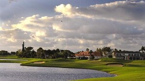 Campo de golf con un estanque, casas al fondo y cielo nublado.