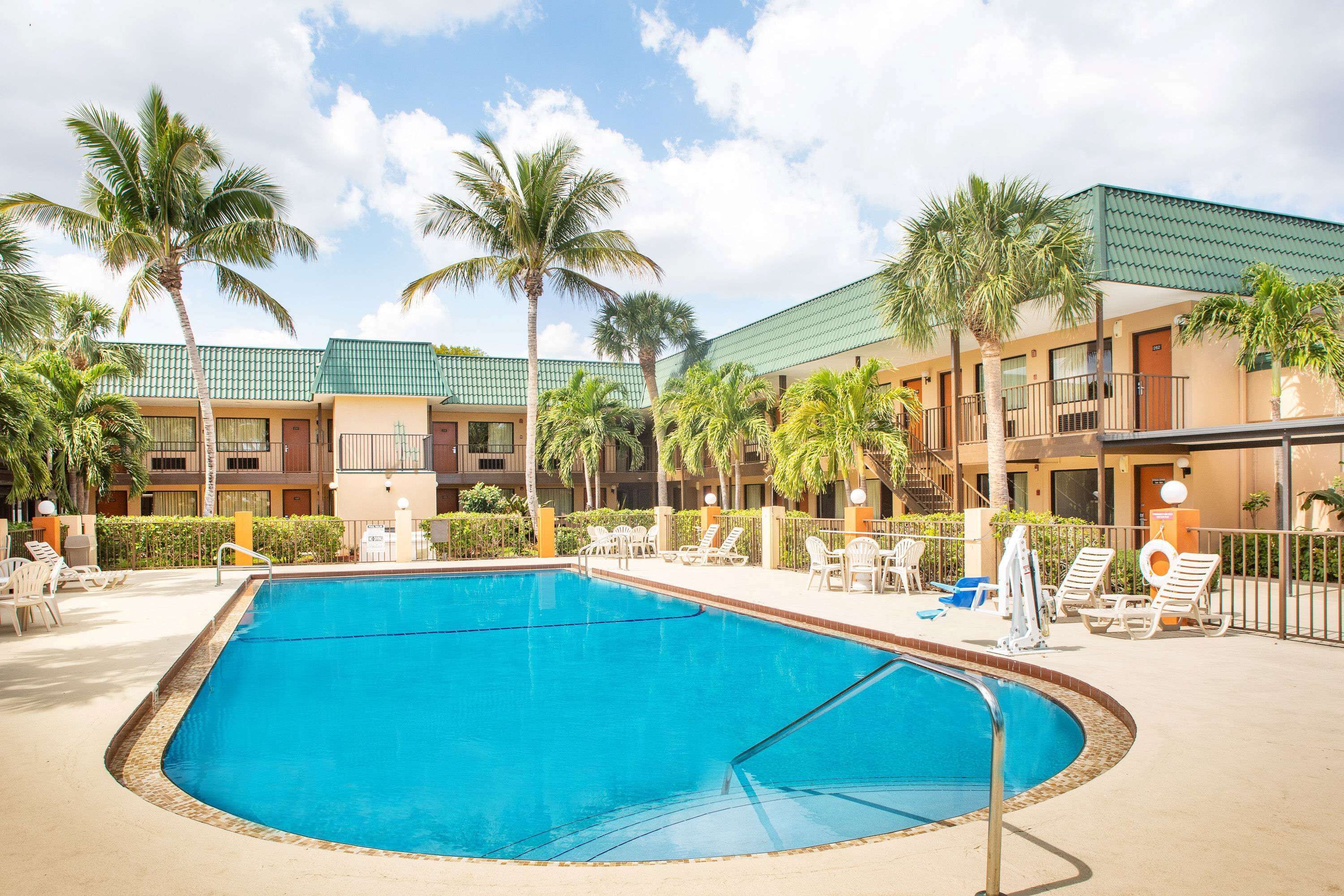 A swimming pool surrounded by lounge chairs and palm trees, with a building featuring balconies and a green roof in the background.