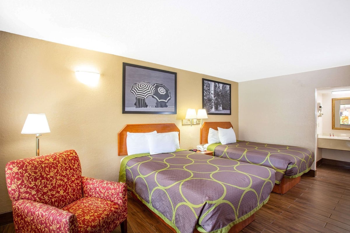 A hotel room with two beds, a chair, and wall-mounted lamps.