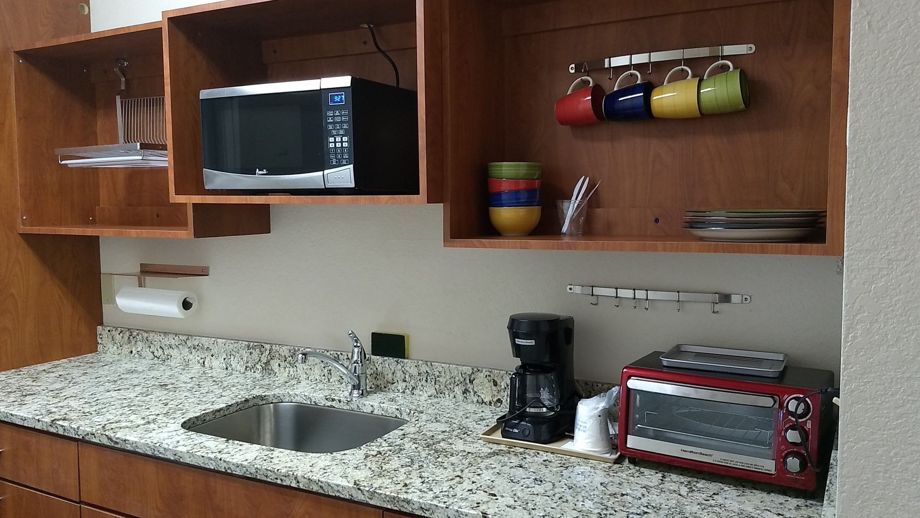 Cocina con encimera de granito, lavamanos de acero inoxidable, microondas, cafetera, tostadora y un expositor con tazas de colores.