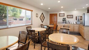 Un espace repas avec des tables et des chaises rondes, un coin cuisine avec des meubles en bois et une fenêtre équipée de stores.