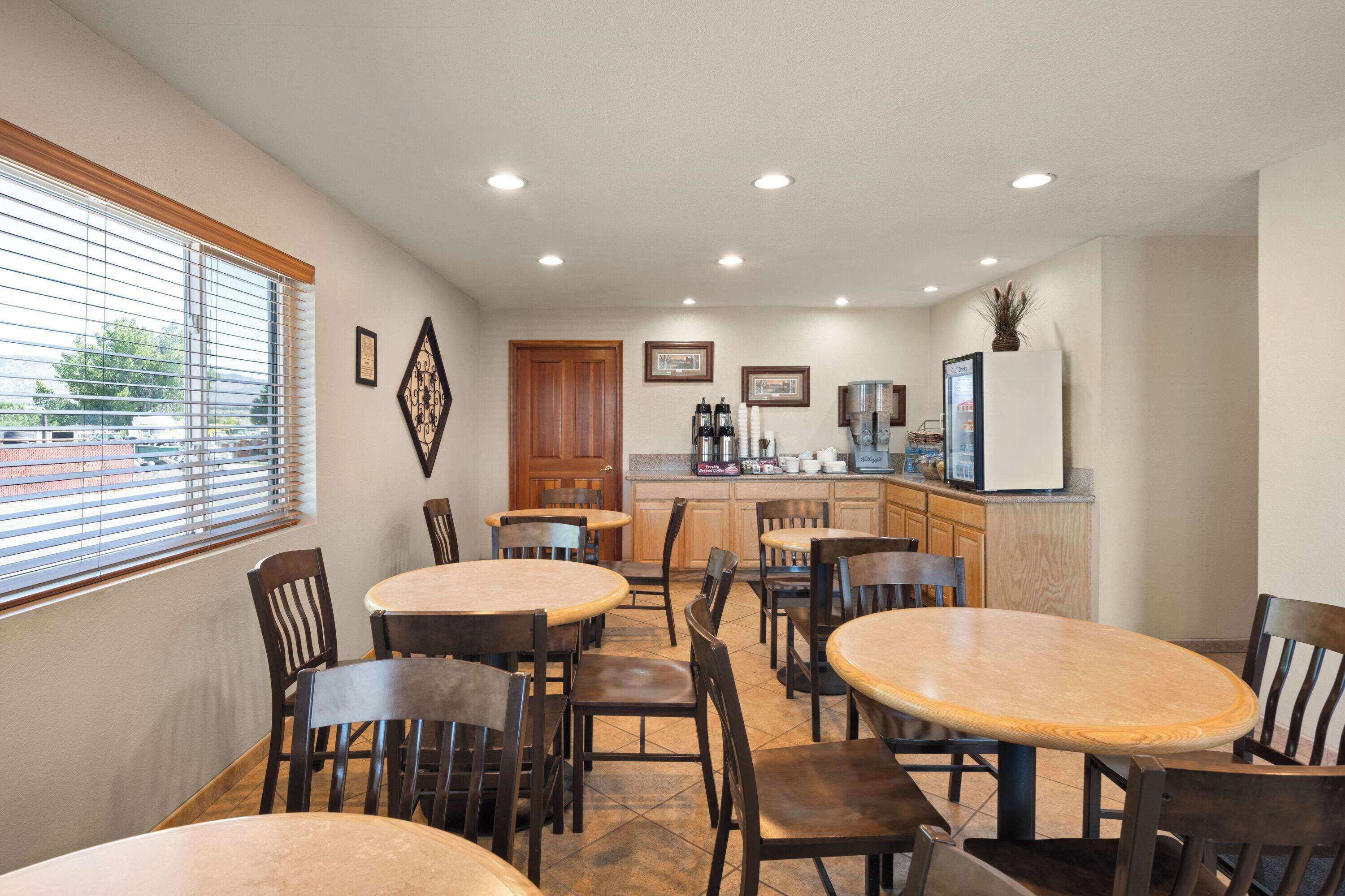 A dining area with round tables and chairs, a kitchen in the background, and a window with blinds.