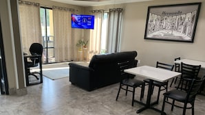 A living room with a black sofa, a dining table with chairs, a TV mounted on the wall, and a framed picture of a cityscape.