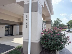 A building entrance with a numbered sign, a covered parking area, and a flower bush.
