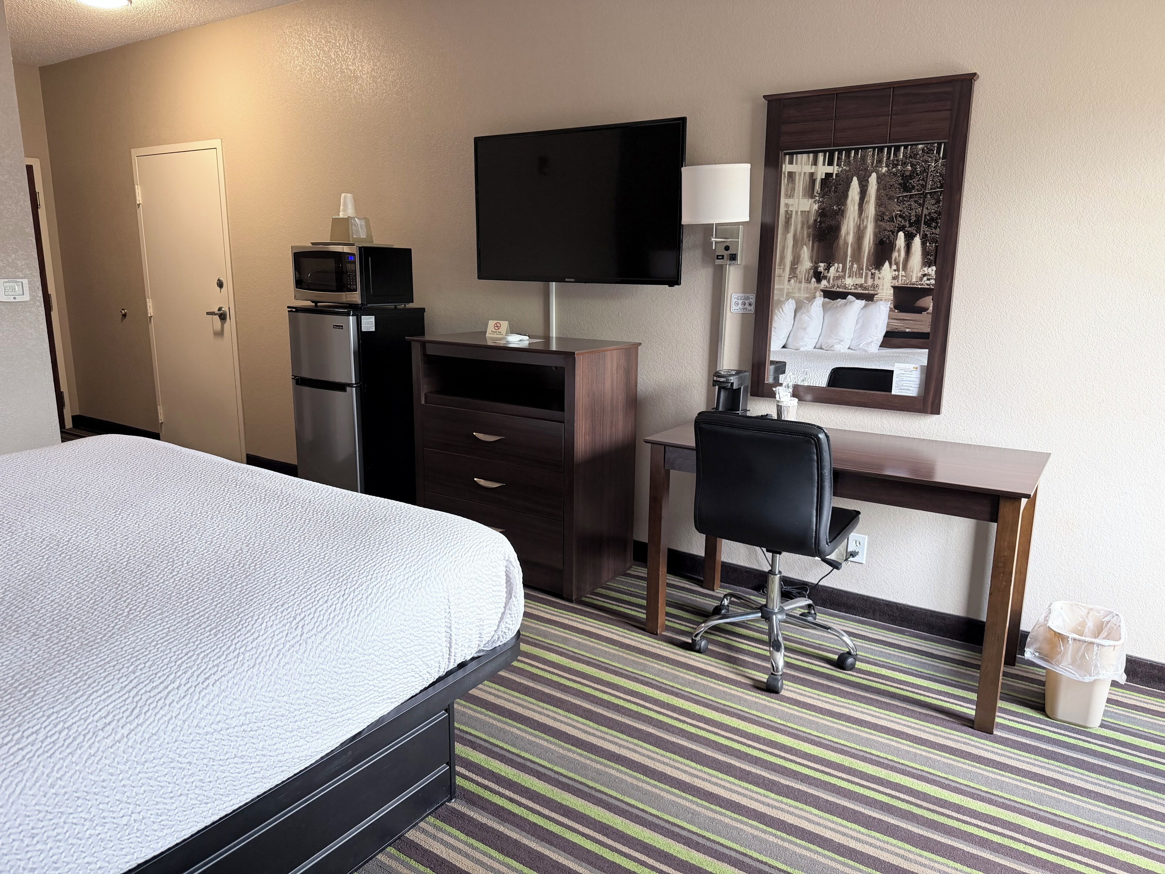 A hotel room with a bed, a desk, a chair, a microwave, a refrigerator, a television, and a mirror.