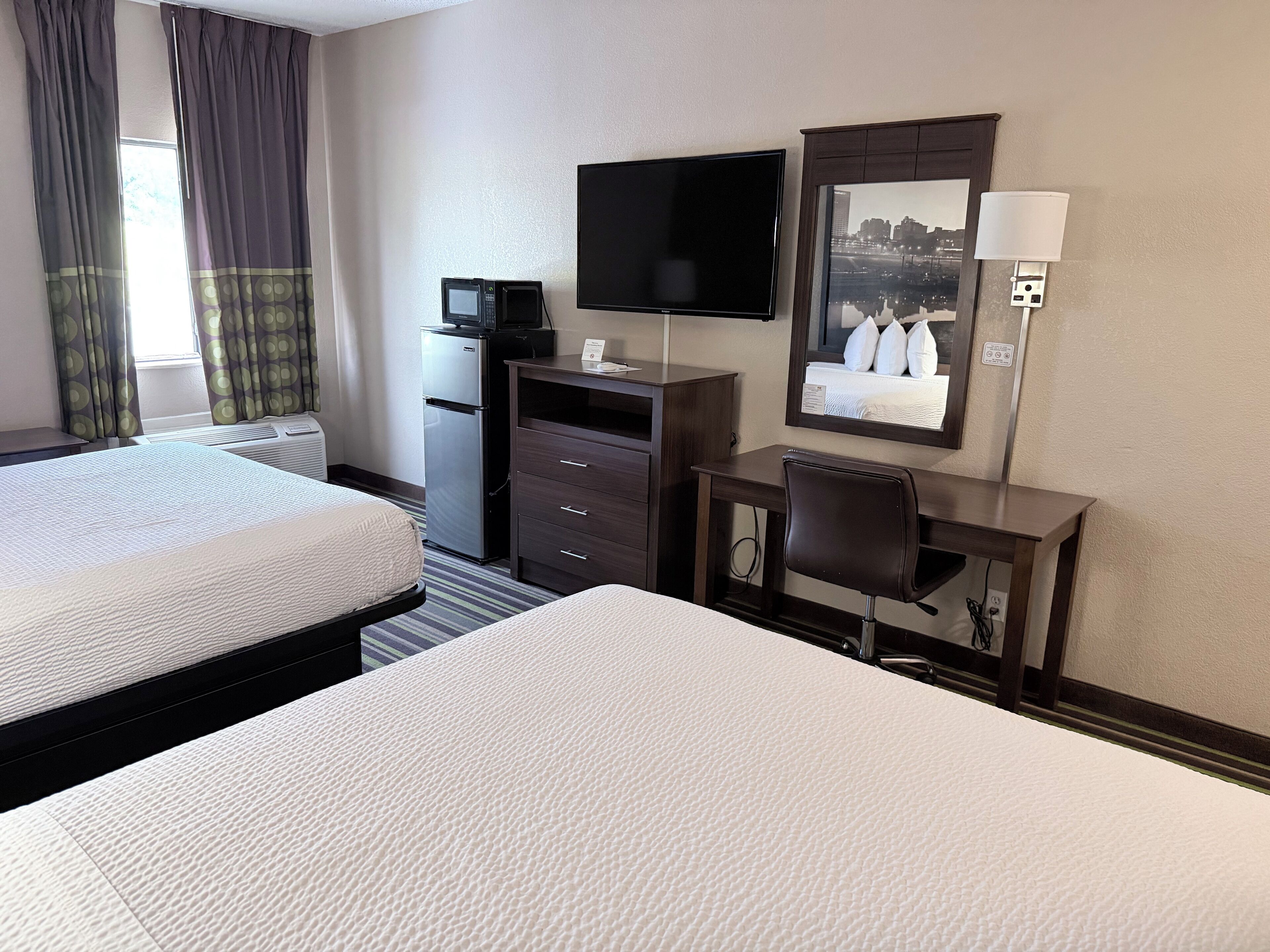 A hotel room with two beds, a desk, a television, and a mirror reflecting a cityscape.