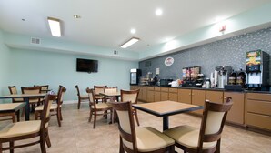 A restaurant with wooden tables and chairs, a tiled floor, a kitchen area with appliances, and a flat-screen TV on the wall.