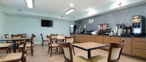 A restaurant with wooden tables and chairs, a tiled floor, a kitchen area with appliances, and a flat-screen TV on the wall.