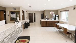 Un vestíbulo de hotel con mostrador de recepción, máquina de café, refrigerador y zona de comedor con sillas y mesa.