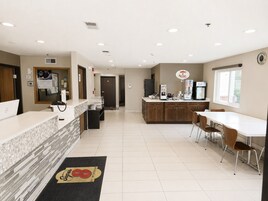 Un vestíbulo de hotel con mostrador de recepción, máquina de café, refrigerador y zona de comedor con sillas y mesa.