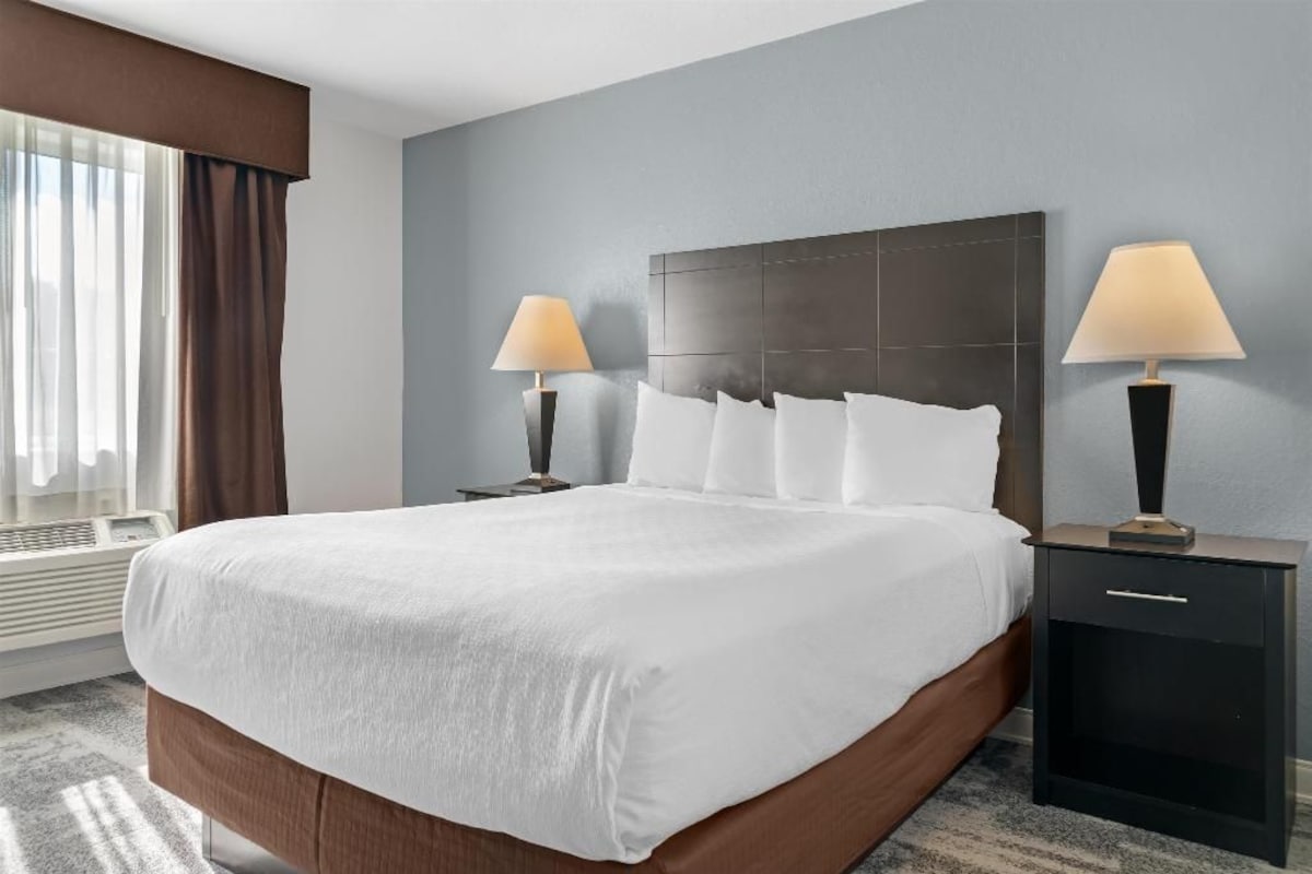 A hotel room with a large bed, two bedside tables with lamps, a window with curtains, and a wall heater.