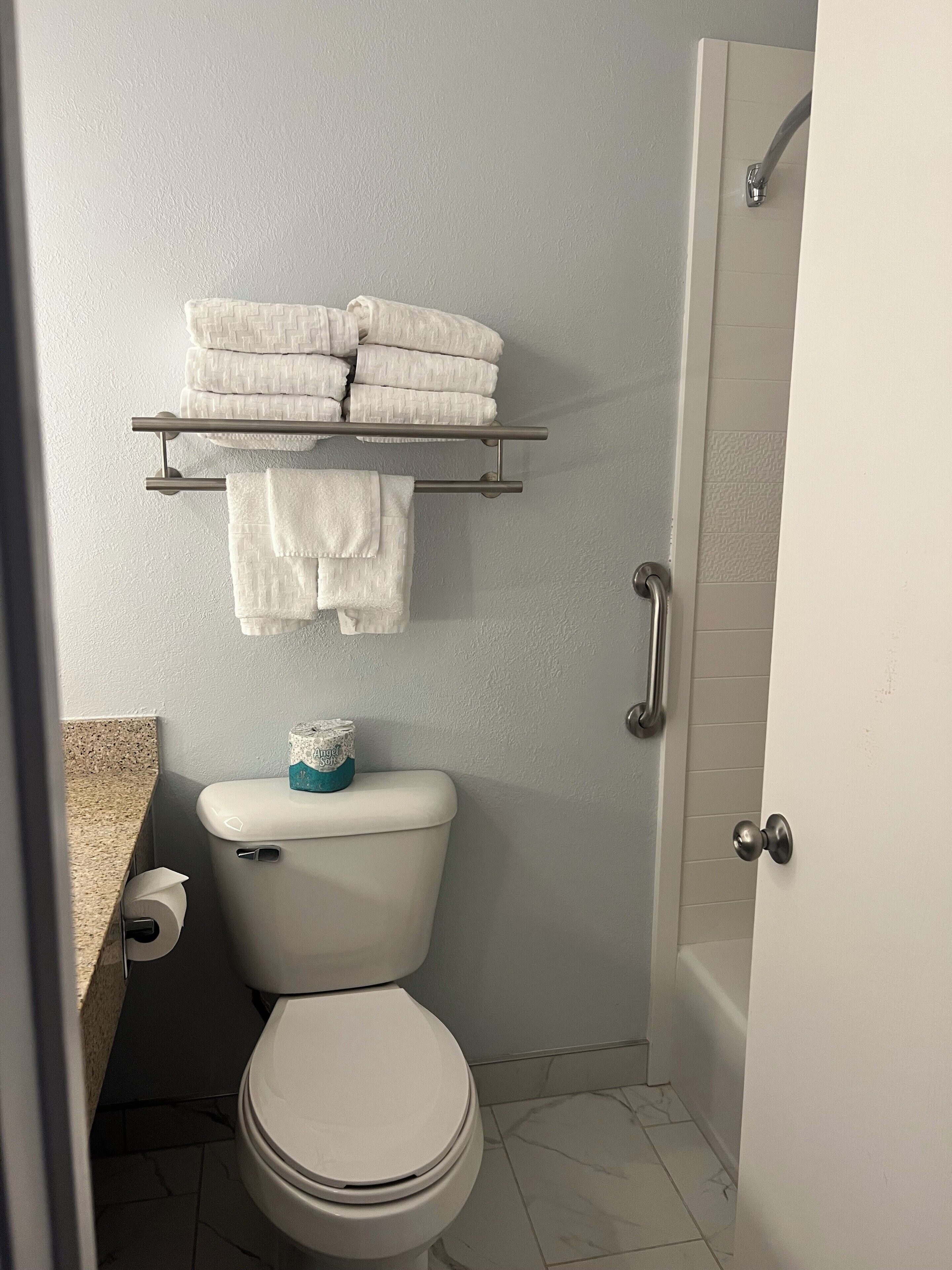 A bathroom with a toilet, a towel rack with folded towels, a toilet paper roll, and a shower door.