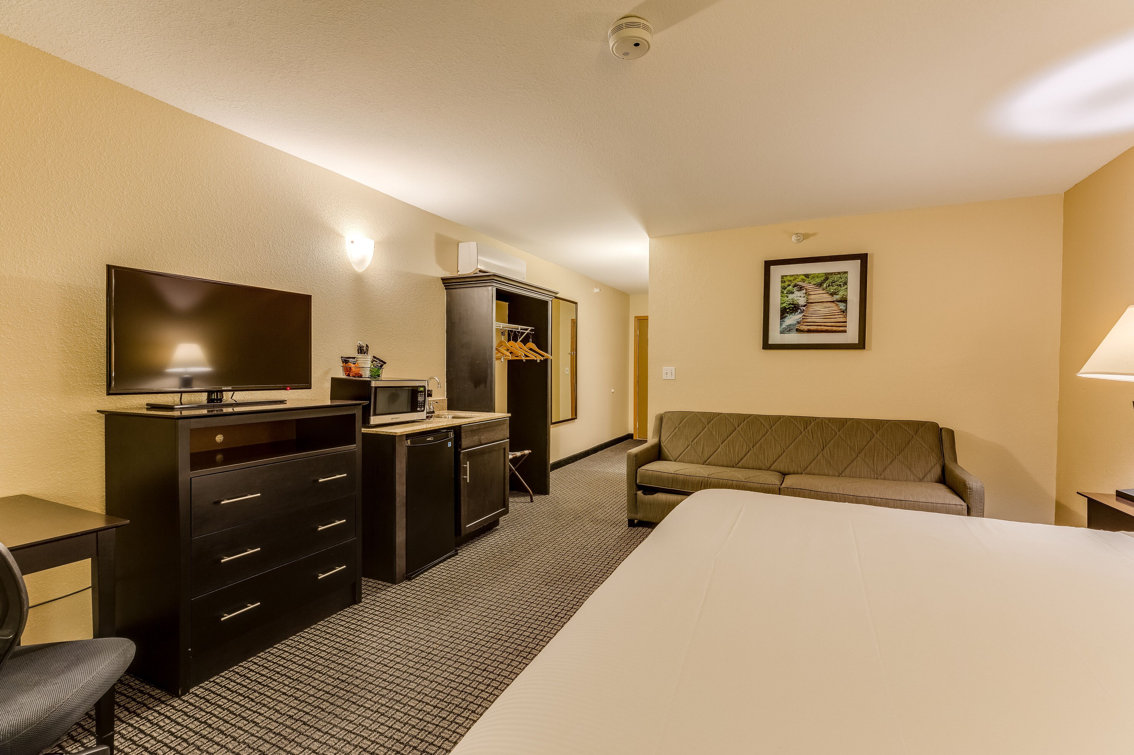 A hotel room with a bed, a TV, a desk with drawers, a microwave, a cabinet, a couch, and a framed picture on the wall.