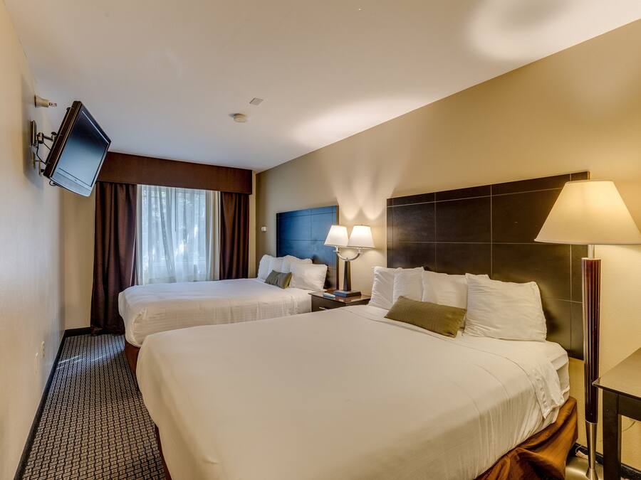 A hotel room with two beds, a flat-screen TV mounted on the wall, a headboard with a built-in lamp, and a small table.