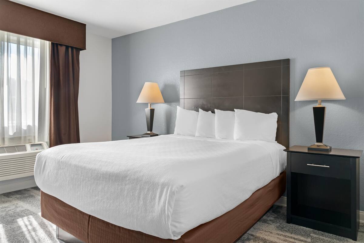 A hotel room with a large bed, two bedside tables with lamps, a window with curtains, and a wall-mounted air conditioning unit.