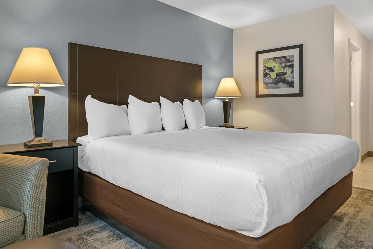 A hotel room with a large bed, two bedside lamps, a headboard, a framed picture on the wall, and a small chair.