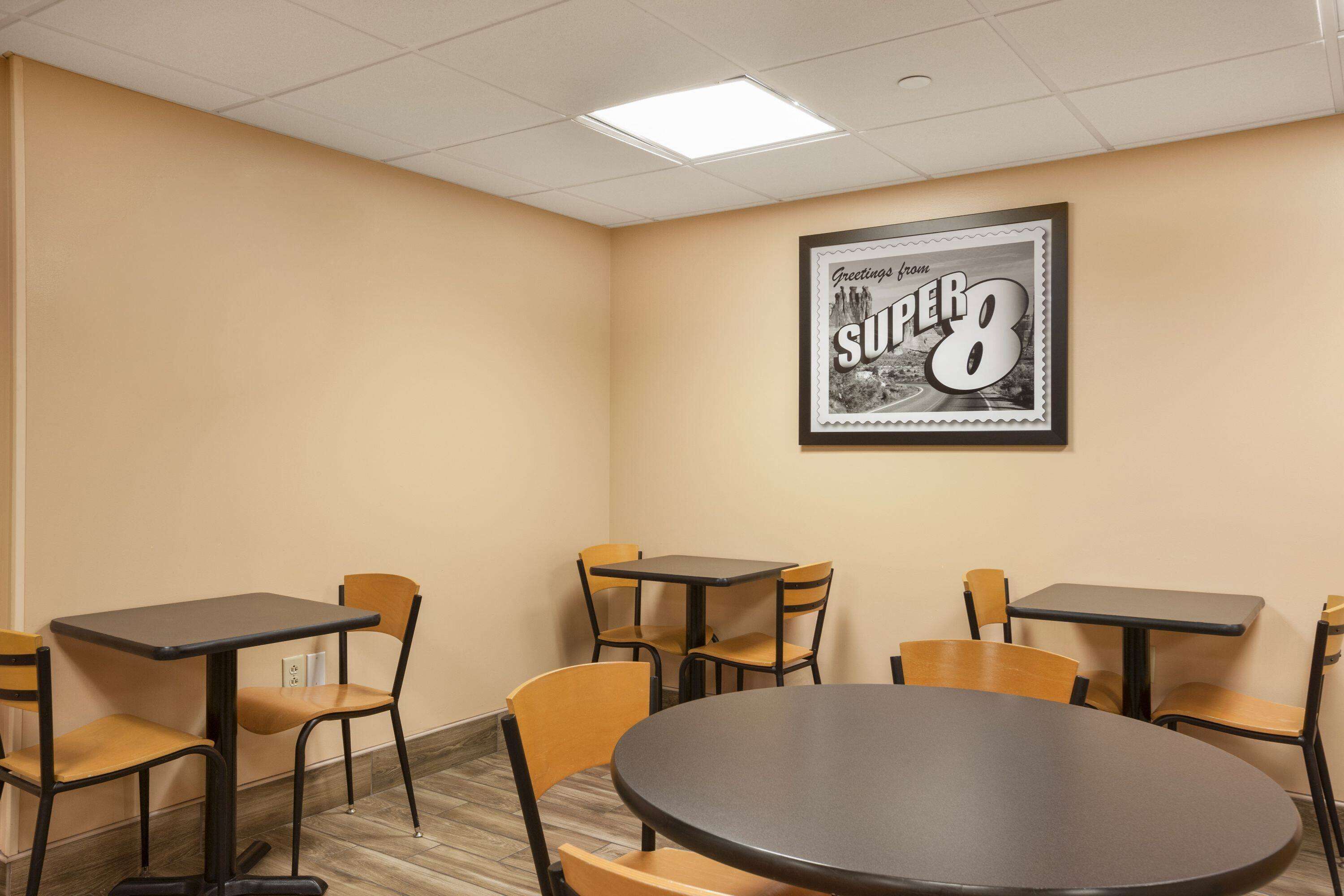 A conference room with a large round table and four rectangular tables, each with two chairs. A framed picture of 'Greetings from Super 8' is on the wall.