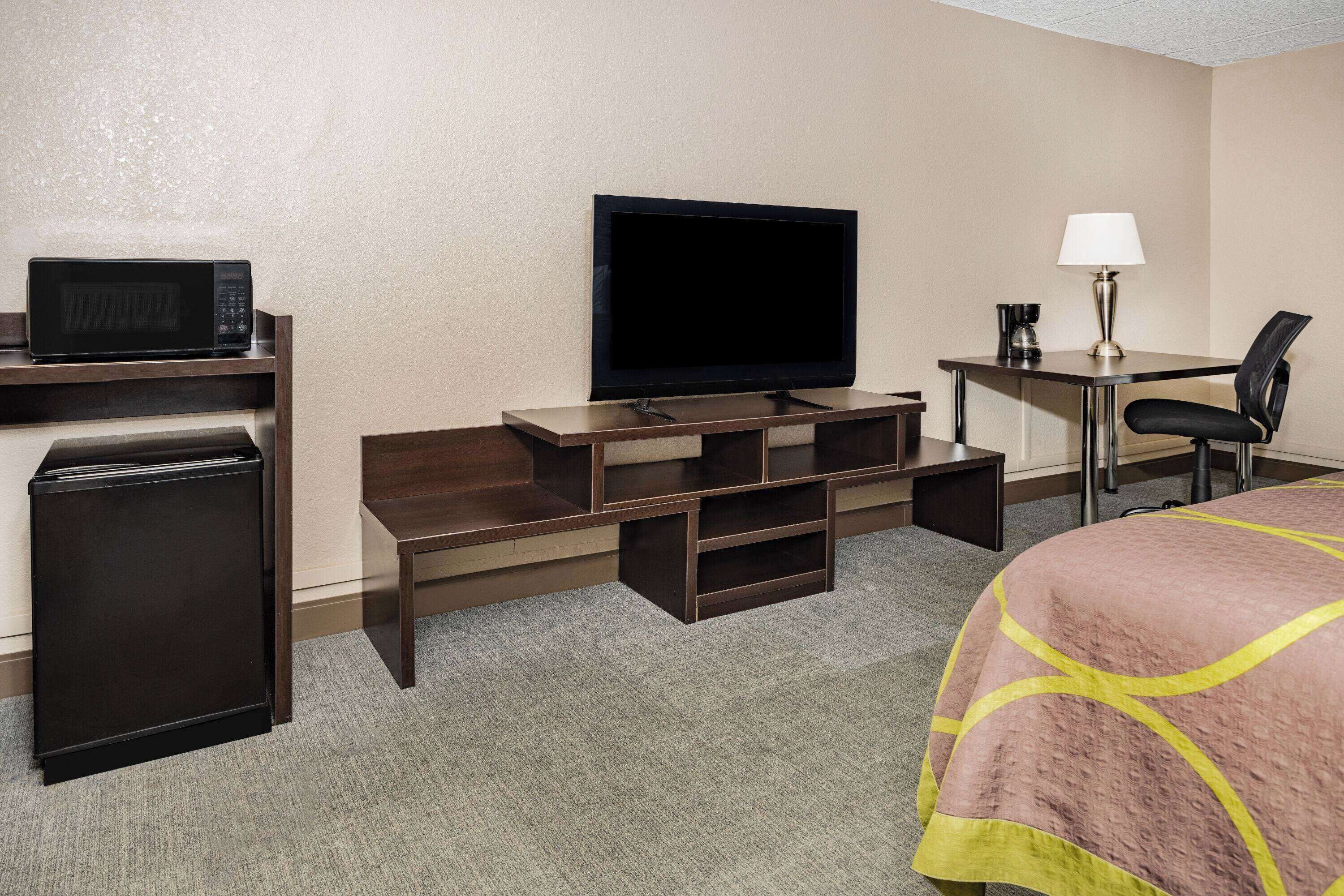 A hotel room with a bed, a desk with a flat-screen TV, a microwave, a refrigerator, and a lamp.