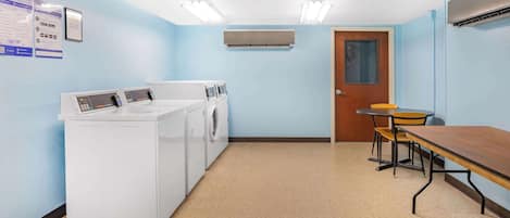 Una lavandería con lavadora, secadora y plancha. Se observa una puerta de madera y una mesa con sillas.