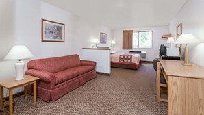 Una habitación de hotel con un sofá rojo, una cama con colcha estampada, un escritorio con lámpara, una mesita y una ventana con cortinas.