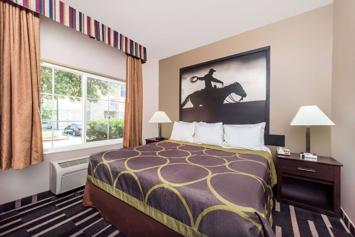 A hotel room with a large bed, two bedside tables with lamps, a window with curtains, a radiator, and a wall-mounted painting of a cowboy on a horse.