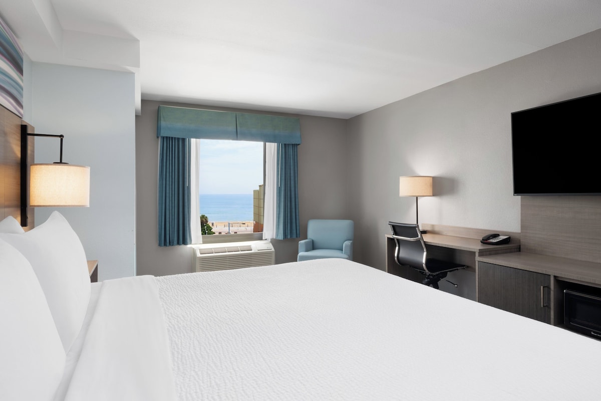 A hotel room with a large bed, a desk, a chair, a TV, and a window with a view of the sea.