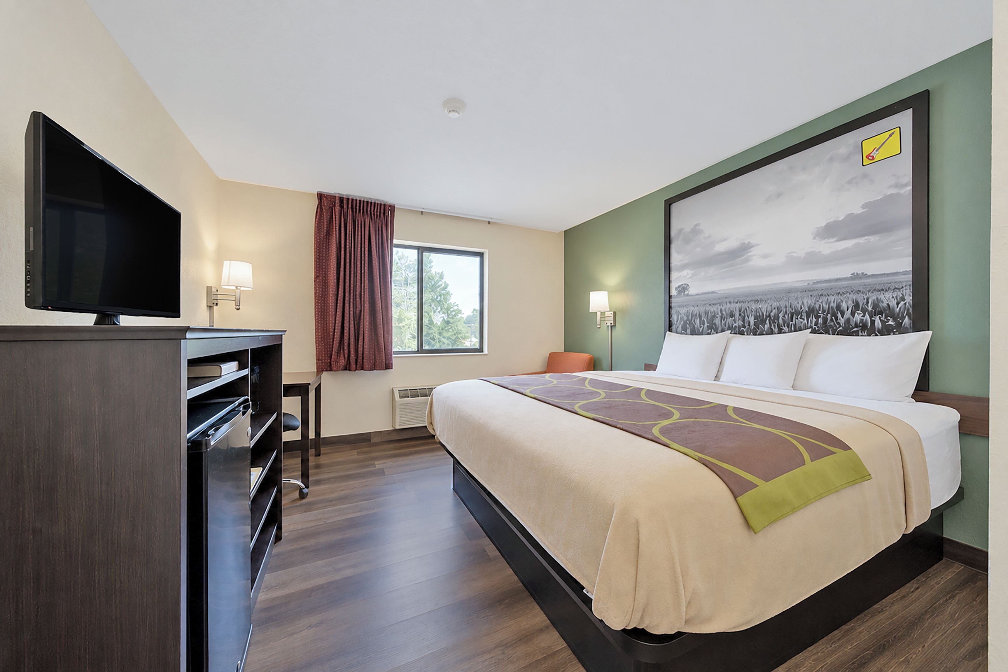 Habitación de hotel con una cama, televisión, escritorio, silla, ventana y un cuadro en la pared.