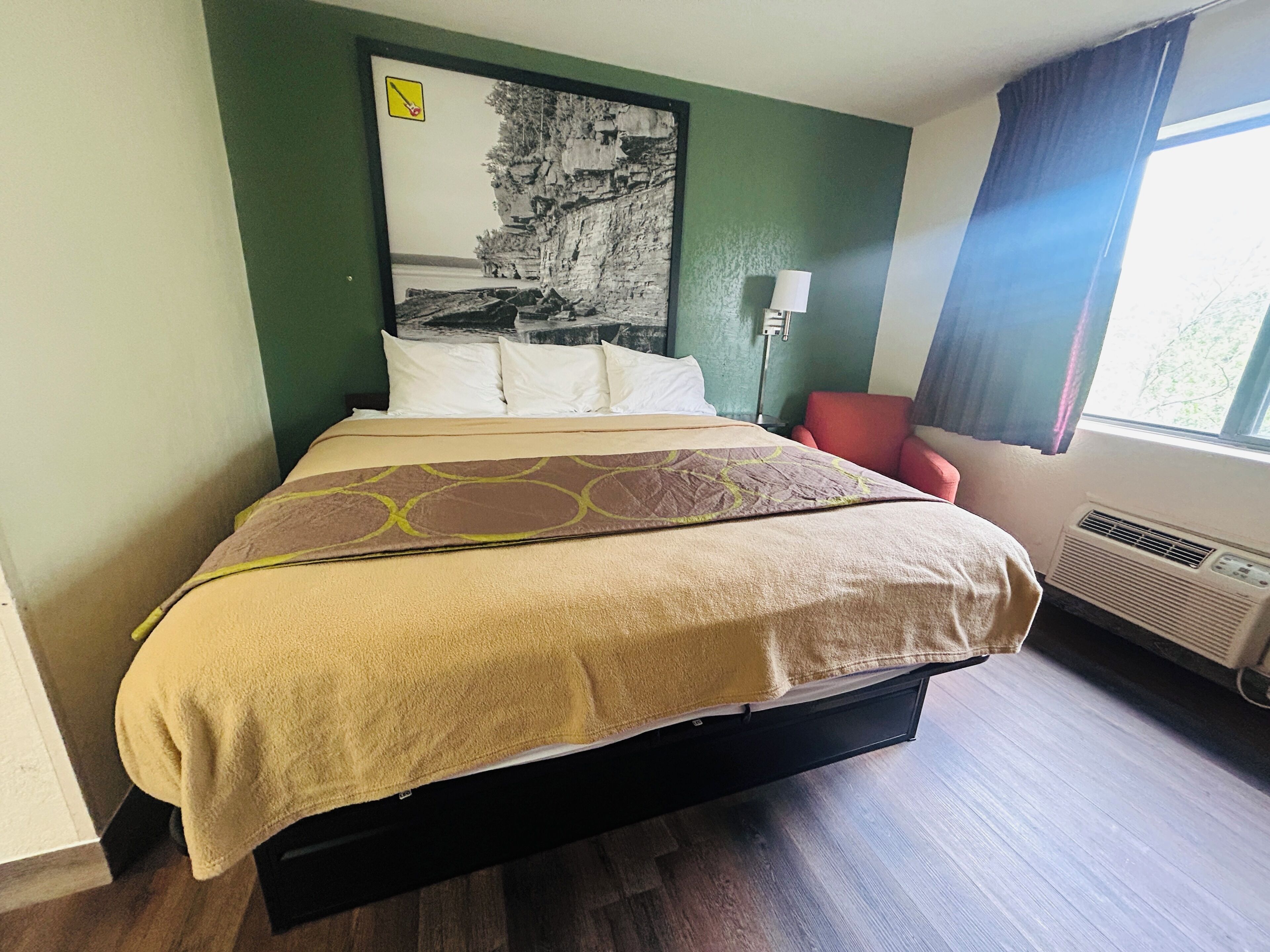 Una habitación de hotel con una cama, una silla roja, una lámpara fijada a la pared y un ventanal con cortinas.