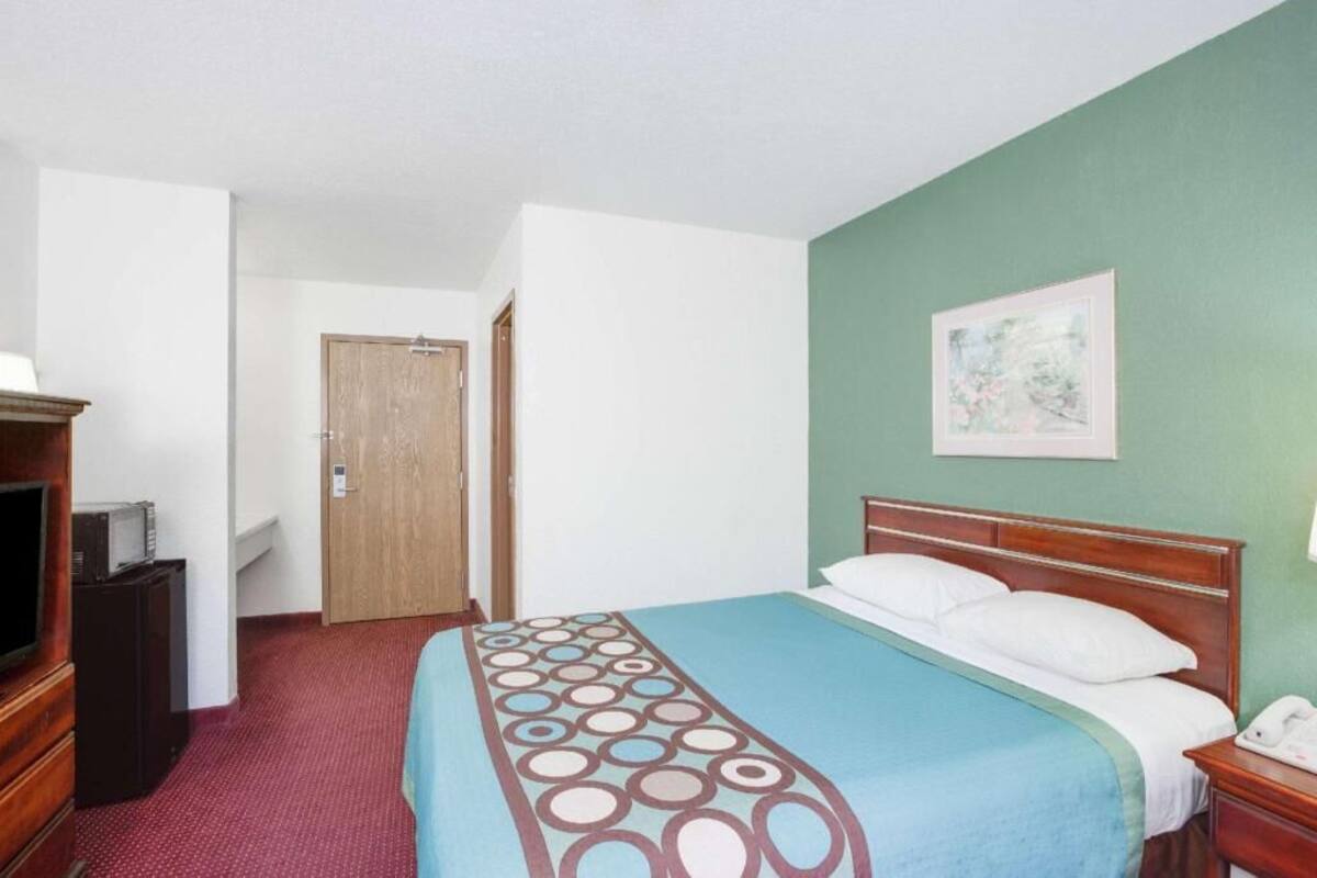 A hotel room with a bed, a wooden headboard, a bedside table with a lamp, a TV, and a door leading to another room.