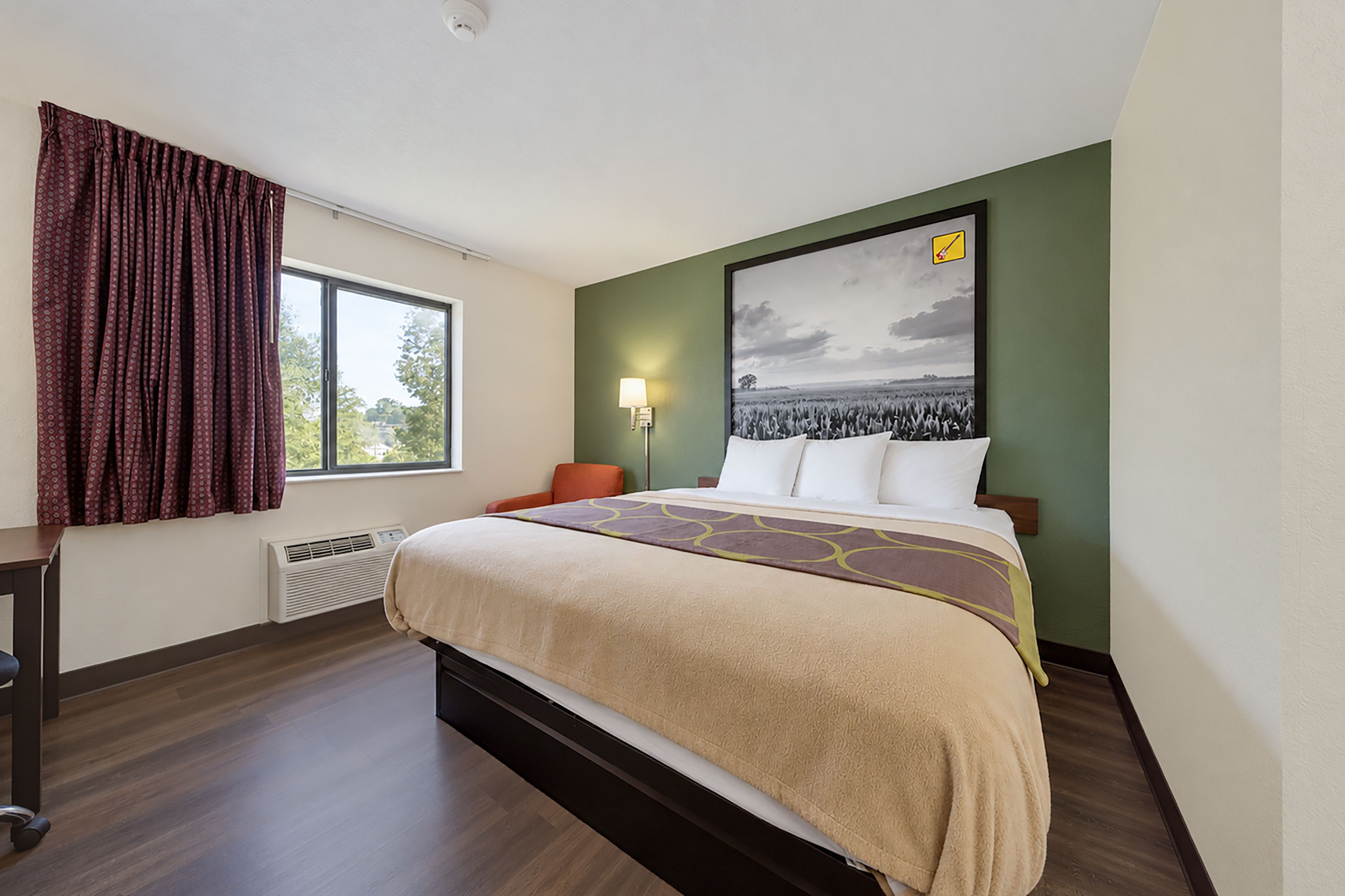 A hotel room with a bed, a window with curtains, a wall-mounted lamp, and a large framed picture.