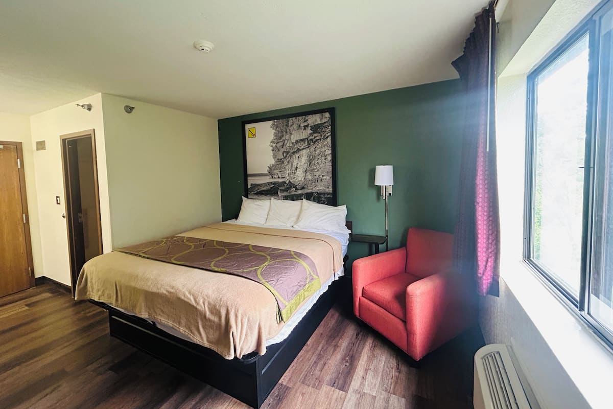 A hotel room with a bed, a red chair, a nightstand, a lamp, and a large window.