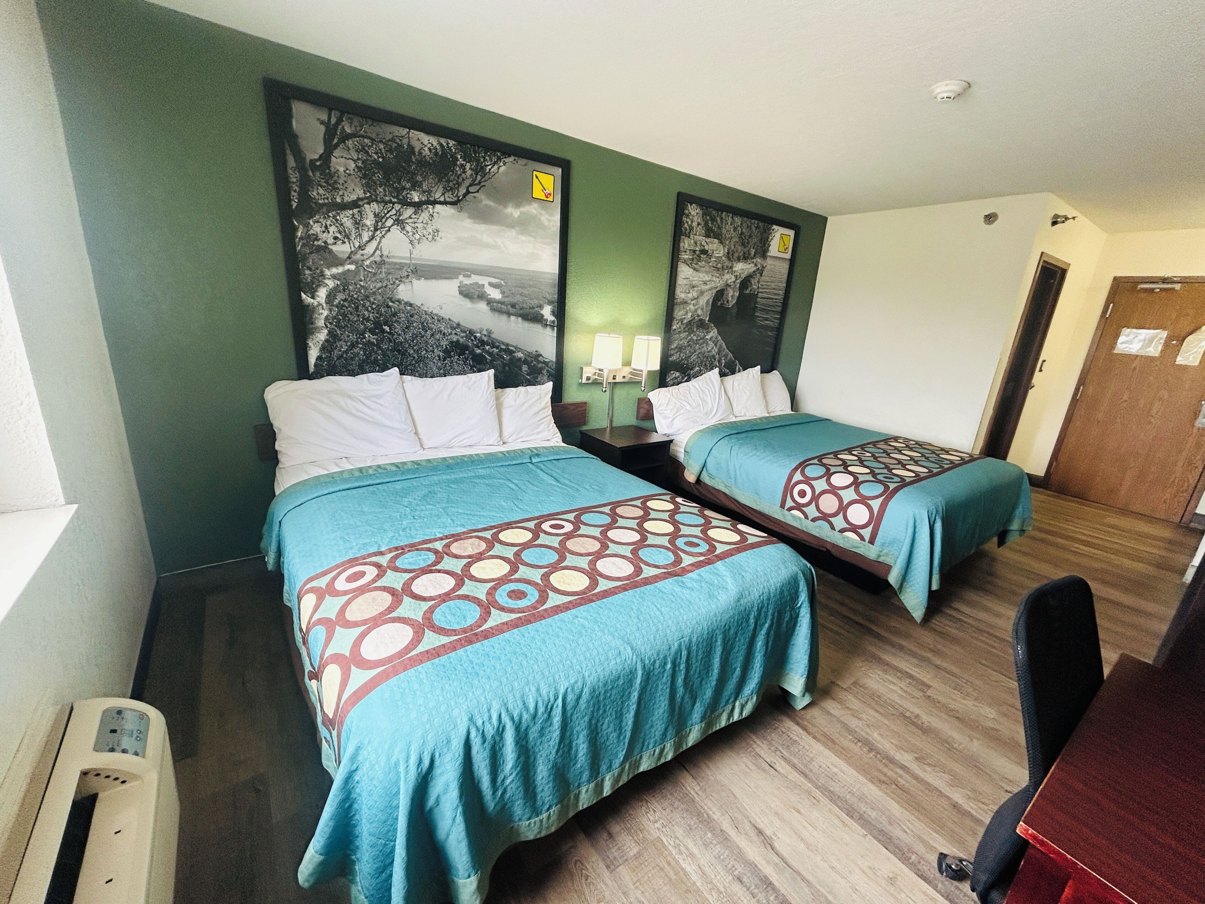 A hotel room with two beds, a desk, and a chair. There are two wall-mounted lamps and a large framed picture above the beds.