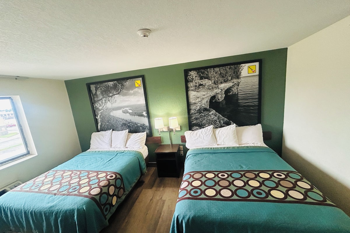 A hotel room with two beds, a window, and two large landscape paintings on the walls.