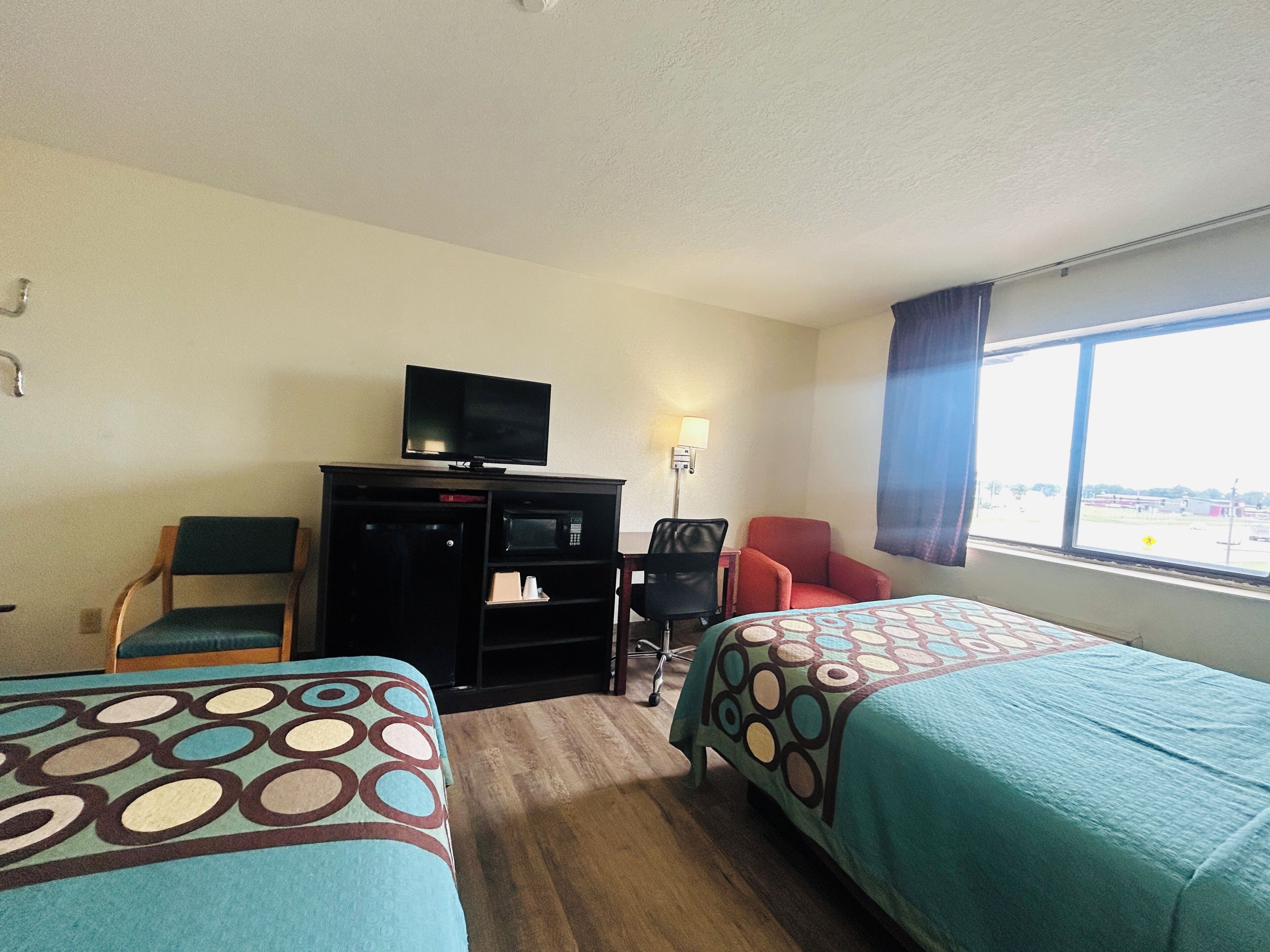 Habitación de hotel con dos camas, un escritorio con televisor, una silla, una lámpara y una ventana con vistas.