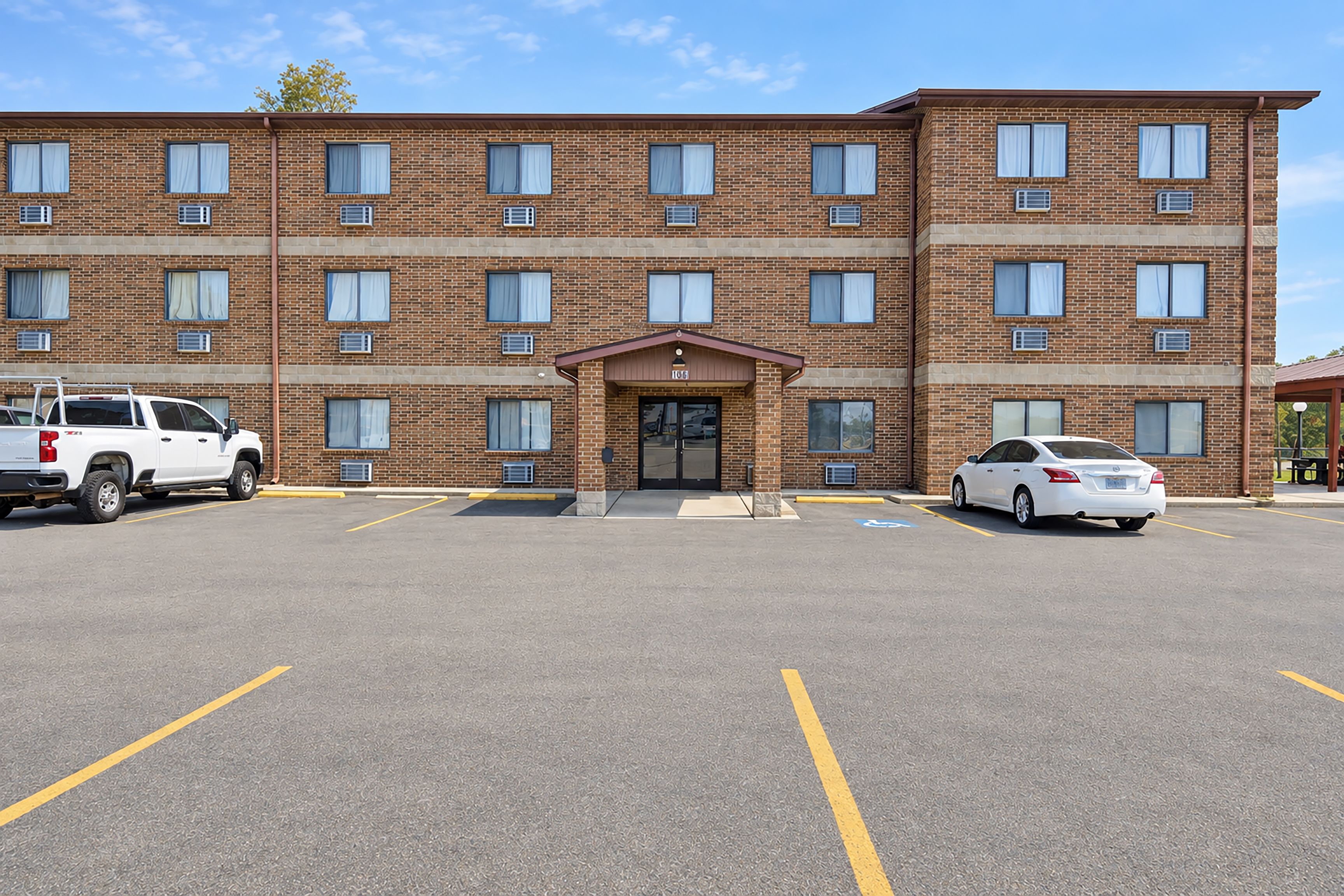 A multi-story brick building with a central entrance, parking lot, and two vehicles parked in front.