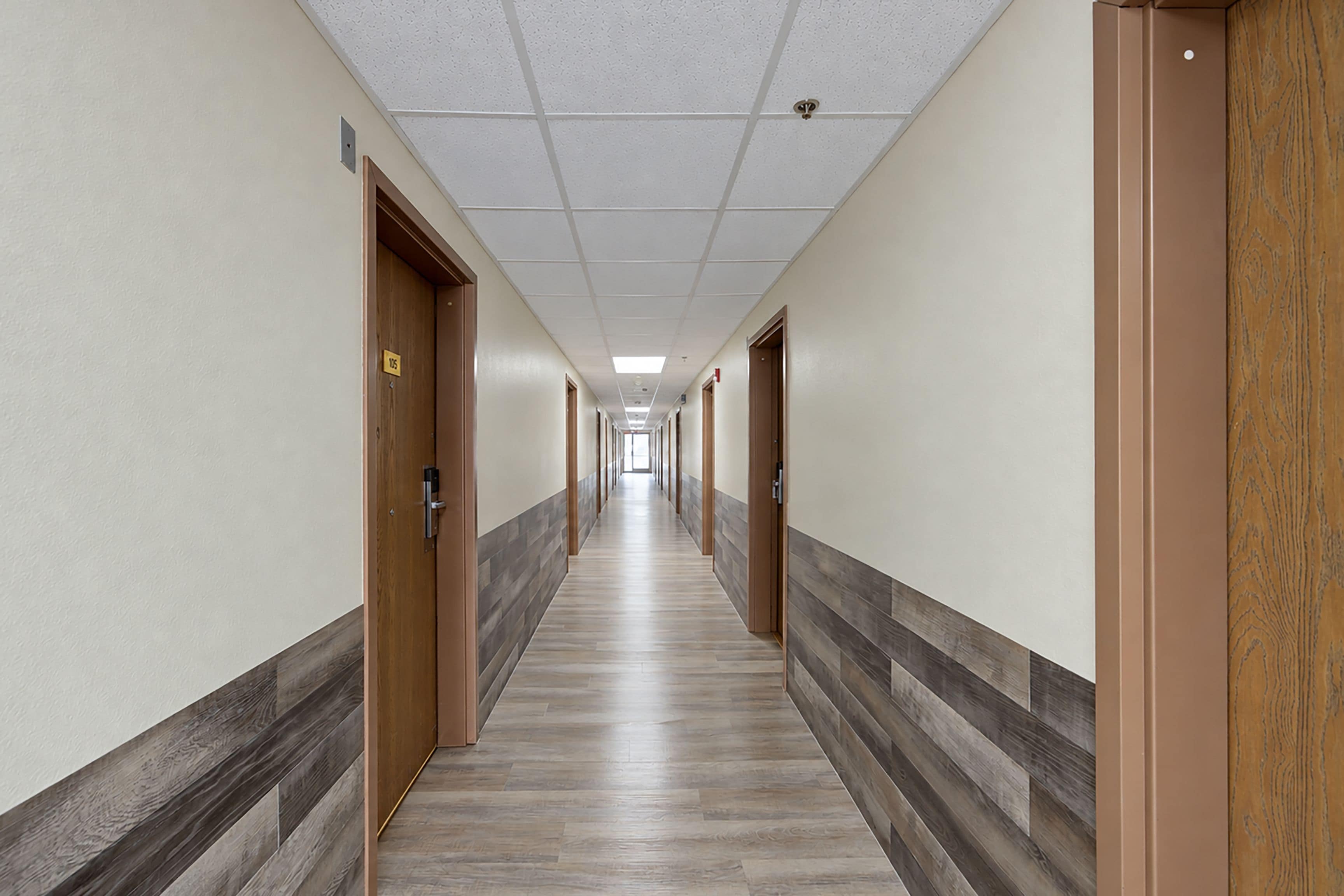 A long, narrow hotel corridor with doors on both sides, a tiled floor, and a ceiling with recessed lighting.