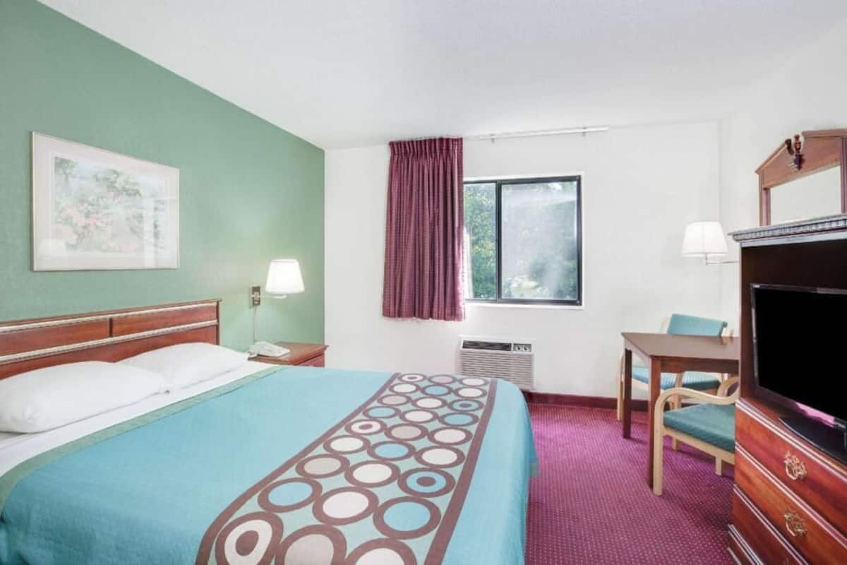 A hotel room with a bed, a desk, a television, a window with curtains, and a wall-mounted picture.
