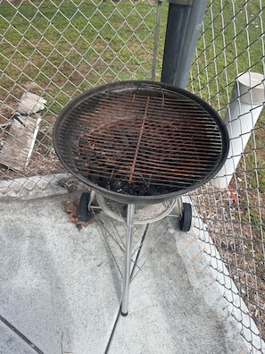A portable grill with a metal grate, set up on a concrete surface, surrounded by a chain-link fence.