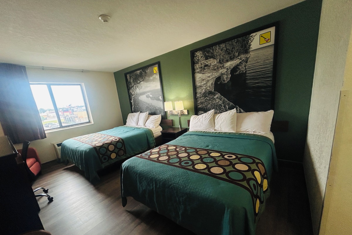 A hotel room with two beds, a desk, a chair, a window with a view, and a painting on the wall.