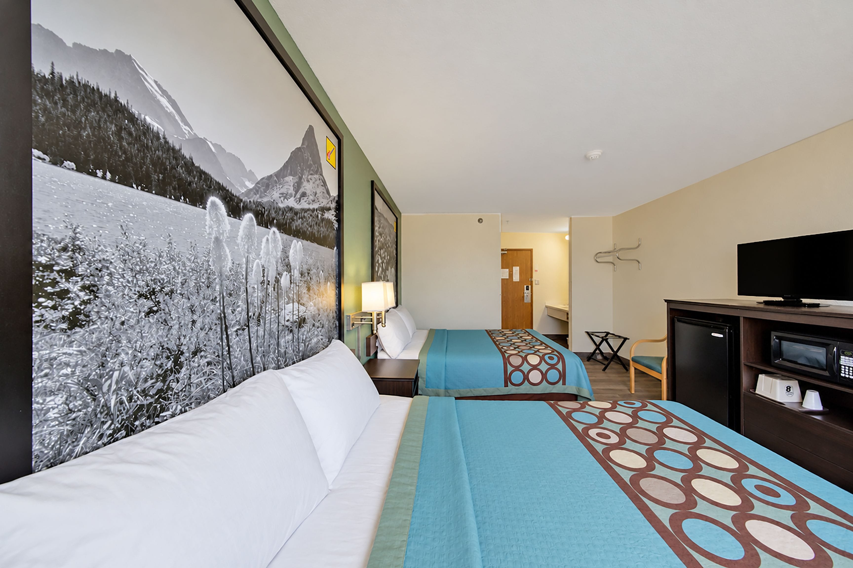 A hotel room with a bed, a desk, a television, and a large mountain view poster on the wall.
