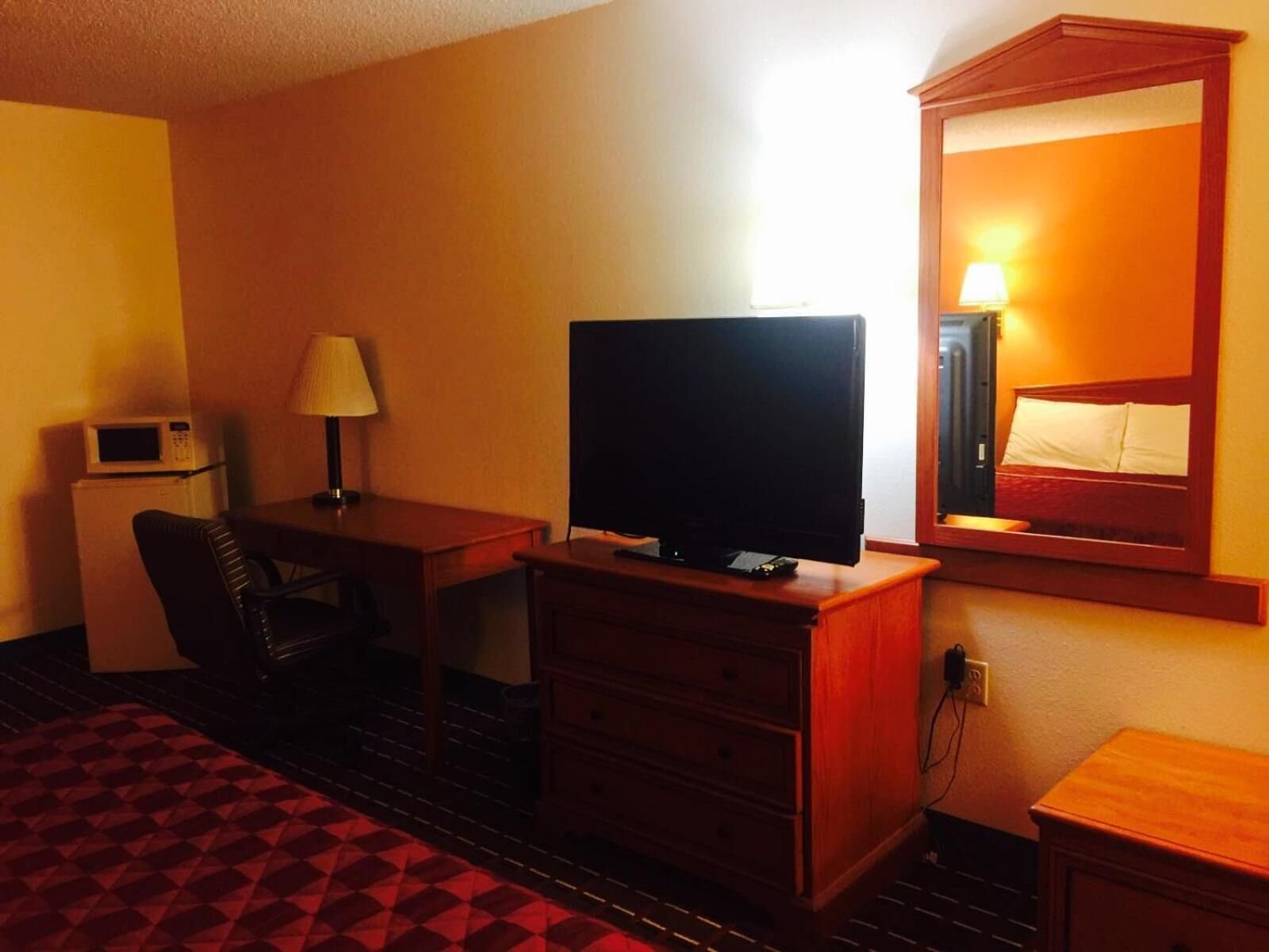 A hotel room with a television on a wooden dresser, a desk with a lamp, a microwave, and a bed with a wooden headboard.